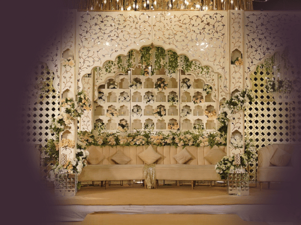 An elaborately decorated stage setup at Nasrullah Marquees, likely for a wedding or formal event. The backdrop features intricate latticework with floral patterns and arches, holding multiple floral arrangements. The stage is adorned with a variety of flowers and greenery, creating a lush and elegant atmosphere. In front of the backdrop, there is a long beige sofa with several matching cushions, flanked by two smaller chairs on either side.