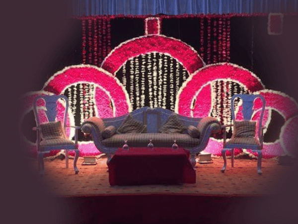 A beautifully decorated event setup at Nasrullah Marquees, featuring a central sofa flanked by two armchairs. The background showcases large, circular floral arrangements in pink and white, adding an elegant and festive touch. In front of the sofa is a small table with decorative items, enhancing the celebratory ambiance.