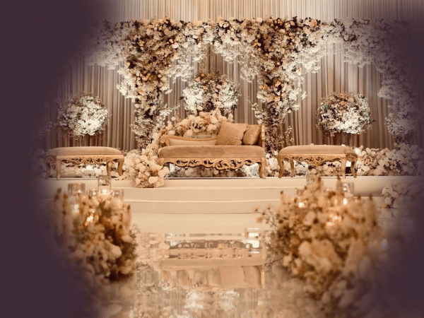 An elaborately decorated stage setup at Nasrullah Marquees, likely for a wedding or a formal event. The stage features a central ornate sofa with cushions, flanked by two smaller matching chairs. The backdrop is adorned with an abundance of flowers and floral arrangements, creating a lush and elegant atmosphere. The floor leading up to the stage is also decorated with flowers and candles, adding to the opulent and romantic ambiance. The overall color scheme is soft and neutral, with shades of white, cream, and gold, enhancing the luxurious feel of the setting.