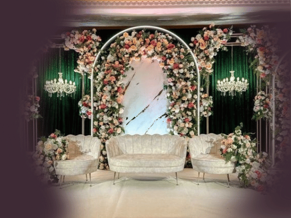 An elegant stage setup at Nasrullah Marquees for a wedding or special event featuring a central arch adorned with lush pink, white, and peach flowers. Flanked by two smaller floral arches, the scene includes green velvet curtains, crystal chandeliers, and a set of three upholstered seats in light colors.