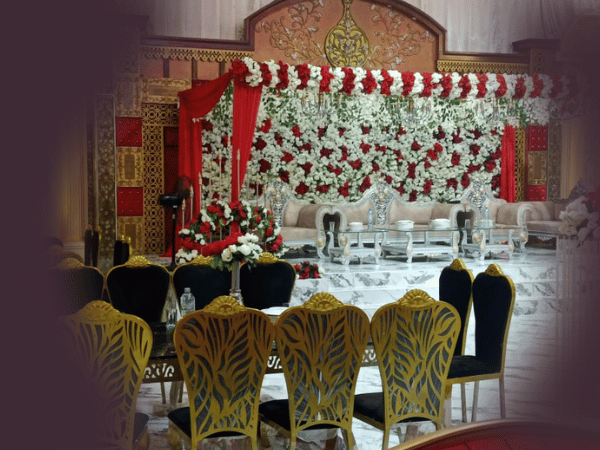 An elaborately decorated stage setup at Nasrullah Marquees for a wedding or formal event. The stage is adorned with a backdrop of red and white flowers, framed by red drapes. Several ornate white and silver chairs are arranged on the stage. In front of the stage, tables and chairs for guests are decorated with floral arrangements. The overall setting is luxurious and elegant, featuring black chairs with gold designs on the backrests.