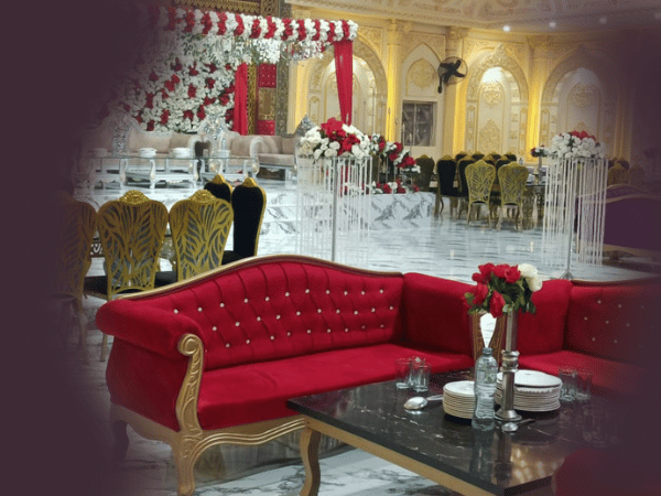 Indoor view of Nasrullah Marquees, showcasing a lavishly decorated banquet hall set up for a wedding or formal event. The hall features golden accents and ornate designs on the walls and ceiling, with a stage at the far end adorned with a floral arrangement of red and white flowers. The foreground displays a red upholstered sofa with golden legs around a black marble table set with plates, glasses, and a vase with red and white flowers. Matching chairs and tables fill the hall, creating a luxurious and festive ambiance.