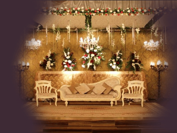 An elaborately decorated stage setup at Nasrullah Marquees for a wedding or formal event. The stage features a luxurious sofa with multiple cushions in the center, flanked by two ornate chairs on either side. The backdrop is adorned with golden fabric and multiple floral arrangements consisting of red, white, and green flowers. Above the stage, hanging floral decorations and chandeliers add to the elegance of the setup. The floor is covered with a patterned rug, and additional light fixtures on either side of the stage contribute to the overall opulent ambiance.