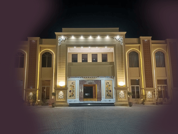 The outdoor view of Nasrullah Marquees at night, showcasing a large, well-lit building with a grand entrance. The facade is illuminated with lights highlighting the architectural details, including large columns and decorative panels. The spacious paved area in front of the building suggests it can accommodate a significant number of guests or vehicles.