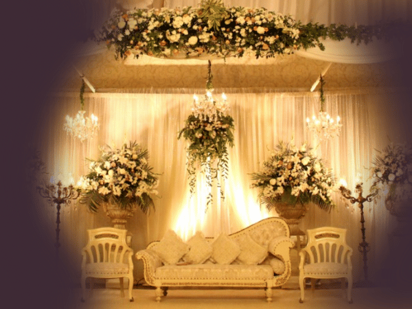 An elegantly decorated stage setup at Nasrullah Marquees, likely for a wedding or formal event. The stage features a luxurious cream-colored sofa with intricate designs and multiple cushions, flanked by two matching chairs. The backdrop consists of draped fabric with soft lighting, creating a warm and inviting ambiance. Large floral arrangements in ornate vases and chandeliers add to the sophisticated atmosphere.