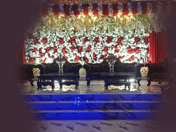 A decorated stage setup at Nasrullah Marquees for a wedding or formal event. The stage features a backdrop of red and white flowers with green leaves, arranged in a dense, patterned manner. There are three ornate, dark-colored sofas with intricate designs on the backrests, placed on the stage. In front of the sofas, there are two small tables with reflective surfaces. The stage itself is made of marble with blue lighting illuminating the steps leading up to it. Above the stage, chandeliers hang, adding to the elegant and festive atmosphere.