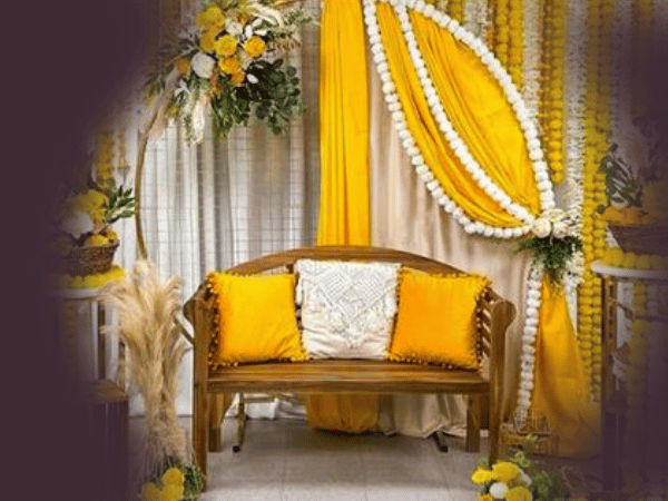 A beautifully decorated event setup at Nasrullah Marquees featuring a wooden bench with three cushions—two yellow and one white. The background displays yellow and white drapery adorned with white garlands and floral arrangements in yellow and white. Additional decorations include a small table with a basket of flowers, a wooden crate with pampas grass, and another table with a basket of flowers, creating a bright and festive atmosphere.